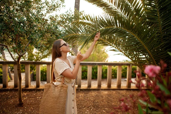 侧视图 黑头发的女人 戴着黑色太阳镜 米黄色的夏装和稻草袋伸出手来的棕榈叶 漂亮的女性在有趣的观光地点散步 — 图库照片