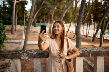 Önde, uzun saçlı, balyalara yaslanan ve cep telefonuyla selfie çeken muhteşem genç bir kadın var. Omzunda saman çantasıyla ağaçların arasında poz veren ve gülümseyen pozitif kadın..