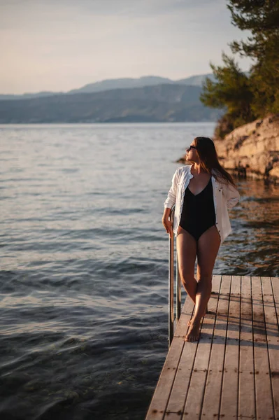 身穿黑色游泳衣和白衬衫的瘦黑发女人站在木制码头上观看日落 在克罗地亚布拉赫岛海滨摆姿势的瘦小女性 — 图库照片