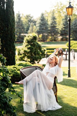 Düğün makyajlı, açık havada sandalyede dinlenen neşeli beyaz bir gelin. Her anın tadını çıkar. Olumlu insan duyguları.