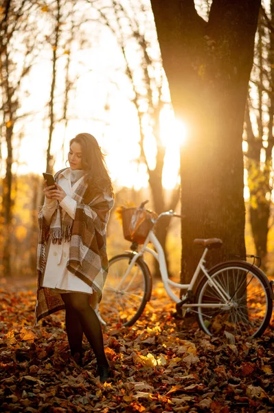 秋天的森林里 一个年轻的微笑的女人 带着一辆自行车在夕阳西下的时候给智能手机发短信 活泼的女孩在大自然中休息 健康生活方式概念 — 图库照片