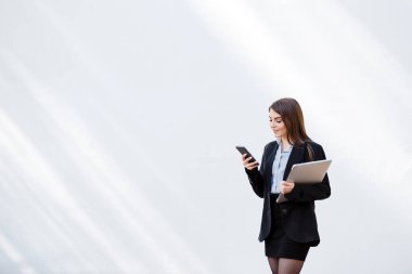 Avrupalı iş kadını cep telefonu kullanıyor. Odaklanmış genç bir kız resmi bir takım elbise giyer ve beyaz arka planda durur. Modern kadın yaşam tarzı. Uzaktan kumanda ve serbest çalışma kavramı. Boşluğu kopyala