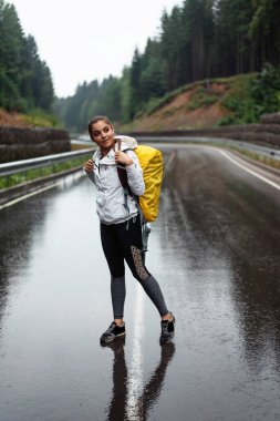 Güzel beyaz kadın yağmurlu havada dağların arasında asfalt yolda duruyor. Yağmurluk giyen ve sırt çantası taşıyan mutlu bir kadın yürüyüşçü..