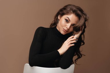 Çekici portresi olan güzel gülümseyen Avrupalı genç bir kadın. Siyah kazak giyen, güzel yüzlü ve saçlı bir kız sandalyeye oturup kameraya bakar. Kahverengi arka planda izole edilmiş. Boşluğu kopyala