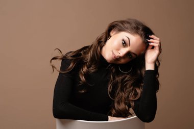 Güzel baştan çıkarıcı genç Avrupalı kadın portresi. Siyah kazak giyen, güzel yüzlü ve saçı olan bir kız sandalyeye oturmuş kameraya bakıyor. Kahverengi arkaplanda izole