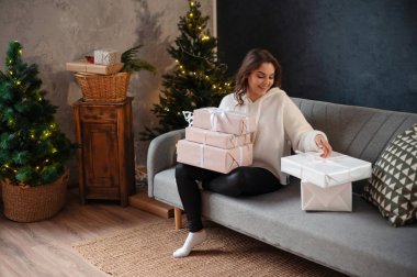 Kıvırcık saçlı mutlu genç bayan Noel ağacının yanındaki kanepede otururken hediyelerini inceliyor. Yeni yıl kavramı