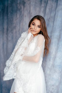 Adorable brunette tender bride in white luxury wedding dress looking at the camera at loft studio interior with blue fabric background 