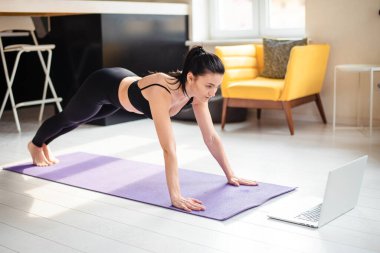Siyah spor elbiseli çekici bir kadın yoga minderinde antrenman yapıyor ve modern laptopta özel ders izliyor. Yurt içi çalışma kavramı.
