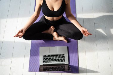 Spor kıyafetleriyle yoga minderinde meditasyon yapan genç bir kadın. Antrenman için modern dizüstü bilgisayar kullanan fitness kadını. Yoga antrenmanını evde yap..