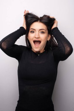 Portrait of amazed beautiful young brunette woman in dark dress standing and looking at camera with surprised face. Indoor studio shot isolated on light white background 