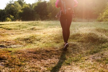 Ormanın yakınındaki yeşil çayırda sırt çantasıyla yürüyen bir kadın. Kızlar spor giyer ve pembe ceket giyer. Dinlenme ve doğa turizmi kavramı. Yaz zamanı. Güneşli gündüz