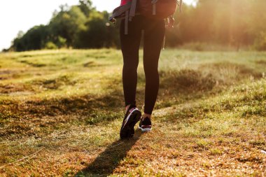 Ormanın yakınındaki yeşil çayırda sırt çantasıyla yürüyen bir kadın. Kızlar spor giyer ve pembe ceket giyer. Dinlenme ve doğa turizmi kavramı. Yaz zamanı. Güneşli gündüz