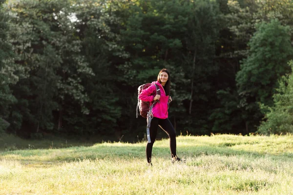 若いヨーロッパの女性の森の近くの緑の牧草地にリュックサックを着て歩く 美しい笑顔の女の子は スポーツウェアとピンクのジャケットを着用 自然の中で休息と観光の概念 晴れた日 — ストック写真