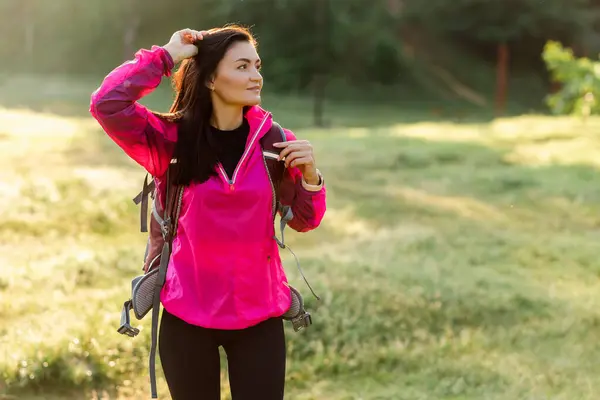 Ormanın yanındaki yeşil çayırda sırt çantasıyla yürüyen Avrupalı genç bir kadın. Çekici odaklı kız spor giyer ve pembe ceket giyer. Dinlenme ve doğa turizmi kavramı. Güneşli gündüz