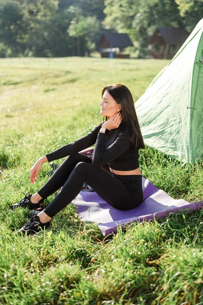 テントの近くのフィットネスマットに座って日光を楽しむヨーロッパのスポーツ少女 緑の牧草地 若い美しい女性はスポーツウェアを着用してください 自然の中で休息と観光の概念 キャンプ休暇だ 晴れた日 — ストック写真