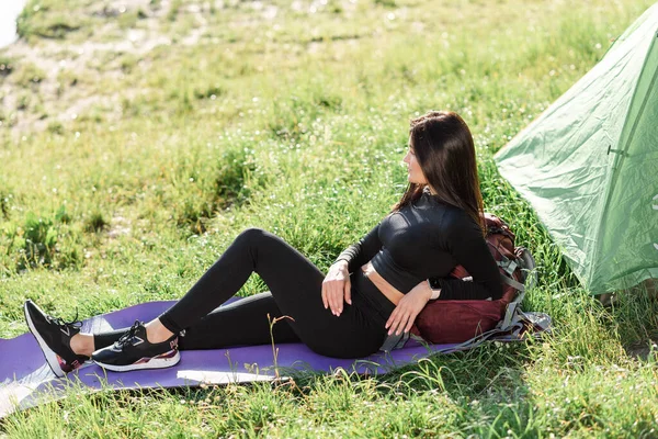 テントの近くのフィットネスマットに横になり 日光を楽しむヨーロッパのスポーツ少女 緑の牧草地 若い美しい女性はスポーツウェアを着用してください 自然の中で休息と観光の概念 キャンプ休暇だ 晴れた日 — ストック写真