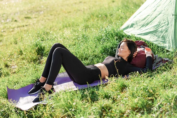 Avrupalı sporcu kız çadırın yanındaki spor minderinde uzanıyor ve güneş ışığının tadını çıkarıyor. Yeşil çayır. Genç ve güzel bir kadın spor giyer. Dinlenme ve doğa turizmi kavramı. Kamp tatili. Güneşli bir gün