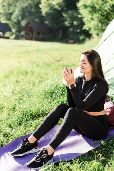 European sport girl sit on fitness mat near tent and drinking tea. Green meadow. Young pretty woman wear sportswear and enjoying sunlight. Concept of resting and tourism on nature. Camping vacation