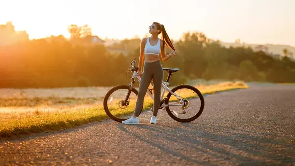 スポーツウェアの魅力的なフィット感とスポーティな女性の肖像画は 日没のフィールド近くの田舎道に自転車で立っています — ストック写真