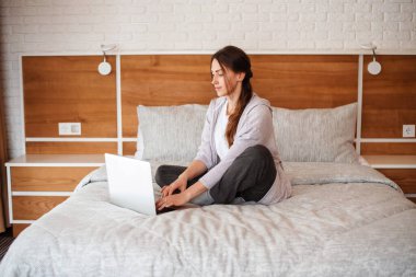 Yatakta oturup dizüstü bilgisayar kullanan güzel Avrupalı kız. Kahverengi saçlı, konsantre olmuş genç bir kadın günlük kıyafetler giyer. Modern apartmandaki yatak odasının içinde. Güneşli gündüz