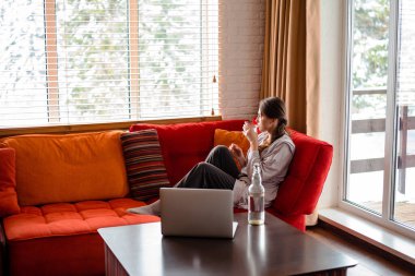 Avrupalı kız evdeki kanepede oturup camdan su içiyor. Genç düşünceli bir kadın günlük kıyafetler giyer. Modern apartmandaki oturma odasının içinde. Dizüstü bilgisayar ve şişe masada.