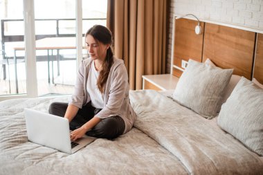Yatakta oturup dizüstü bilgisayar kullanan güzel Avrupalı kız. Kahverengi saçlı, konsantre olmuş genç bir kadın günlük kıyafetler giyer. Modern apartmandaki yatak odasının içinde. Güneşli gündüz