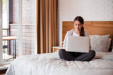 Yatakta oturup dizüstü bilgisayar kullanan güzel Avrupalı kız. Kahverengi saçlı, odaklanmış genç bir kadın günlük kıyafetler giyer. Modern apartmandaki yatak odasının içinde. Güneşli gündüz
