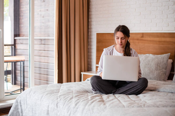 Beautiful european girl sitting on bed and using laptop computer. Young focused woman with brown hair wear casual clothes. Interior of bedroom in modern apartment. Sunny daytime
