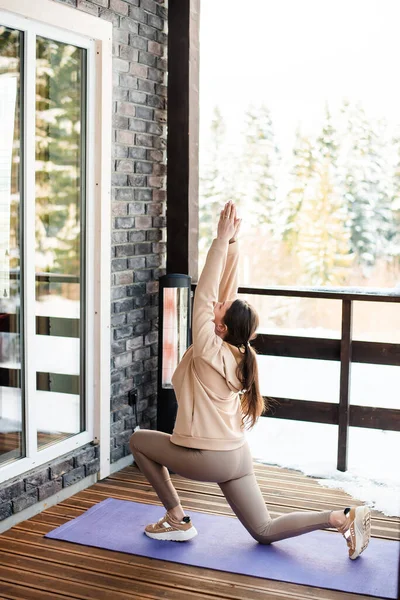 活跃的年轻女子在木制小屋的露天平台上做瑜伽垫上的健身锻炼 背景上的雪地森林 冬季在室外训练 — 图库照片
