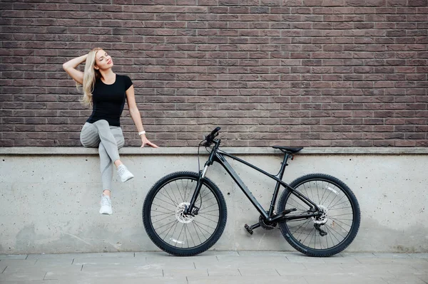 朝のワークアウトの後に休んでいる間 黒い自転車の近くの通りに座って金髪の髪を持つ美しい白人女性 屋外活動後のリラクゼーション サイクリングのコンセプト — ストック写真
