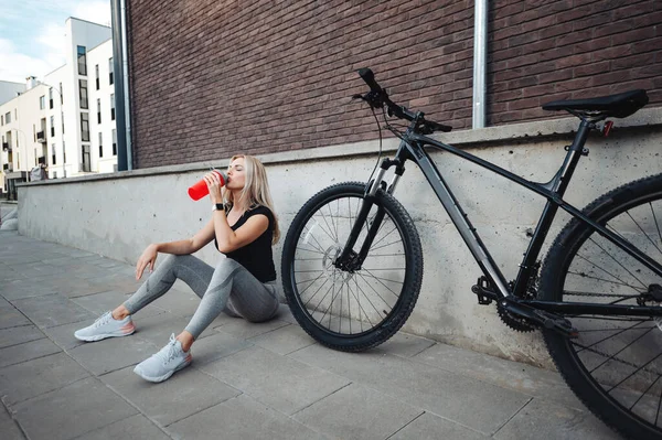 アウトドアで座ってスポーツボトルから水を飲むアクティブウェアで魅力的な白人女性 朝のサイクリングの後に若いブロンドがさわやかな 近くに立つ黒い自転車 — ストック写真