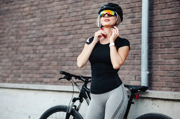Siyah bisikletin yanında duran ve gözlüklü kask takan beyaz kadın. Spor sarışın temiz havada bisiklet sürerken güvenlik ekipmanı kullanıyor..