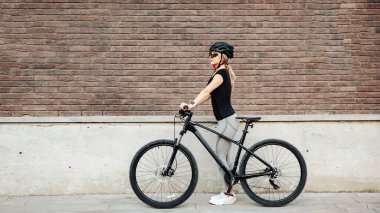 Aktif giyimli, kasklı ve gözlüklü beyaz kadın bisikletçinin şehir caddesinde duran siyah bisikletinin yan görüntüsü. Antrenmandan sonra gevşeme.