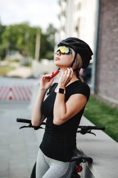 Siyah bisikletle caddede dikilirken güvenlik kaskı takan ve aynalı gözlük takan güzel genç bir kadının yan görüntüsü. Dişi bisikletçi dışarıda antremana hazırlanıyor..