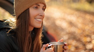 Güzel bayan arabanın bagajında oturuyor ve çay ya da kahve içiyor. Arka planda sonbahar doğası, renkli akçaağaç yaprakları. Macera yolculuğu. Gezgin yaşam tarzı 