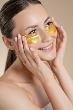 Gözlerinin altında kolajen altın pedlerle bej rengi stüdyo arkasında poz veren pozitif genç bayan. Güzel model güzellik prosedürlerini seviyor. Cilt bakımı kavramı.