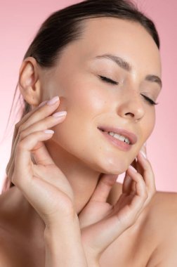 Stüdyoda poz veren, doğal, temiz tenli, şehvetli genç bir bayan. Kahverengi saçlı, gözlerini kapalı ve yanaklarına dokunan beyaz bir kadın. Pembe arka planda izole edilmiş.