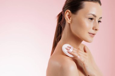 Yanaklarına pudra sürmek için yumuşak sünger kullanan çıplak omuzlu çarpıcı genç bir kadın. Beyaz kadın model hafif makyaj yapıyor. Pembe stüdyo arka planında izole edilmiş..