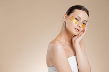 Çıplak omuzlu narin genç bir kadın gözlerinin altında altın bantlar kullanıyor. Bej renkli stüdyo arka planında izole edilmiş. Güzellik ve cilt bakımı.