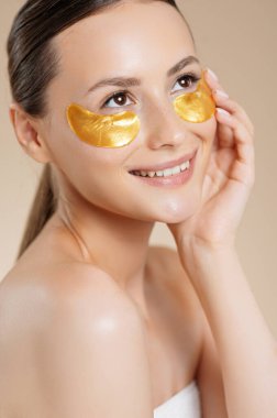 Gözlerinin altında kolajen altın pedlerle bej rengi stüdyo arkasında poz veren pozitif genç bayan. Güzel model güzellik prosedürlerini seviyor. Cilt bakımı kavramı.