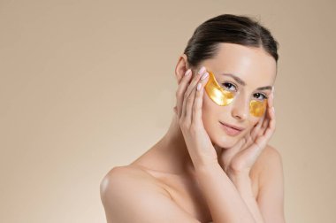 Çıplak omuzlu narin genç bir kadın gözlerinin altında altın bantlar kullanıyor. Bej renkli stüdyo arka planında izole edilmiş. Güzellik ve cilt bakımı.