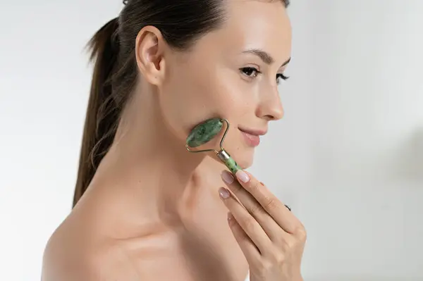 Sensual young woman massaging face with jade roller over white studio background. Caucasian model taking care of natural beauty. Beauty routine and skin procedures concept.