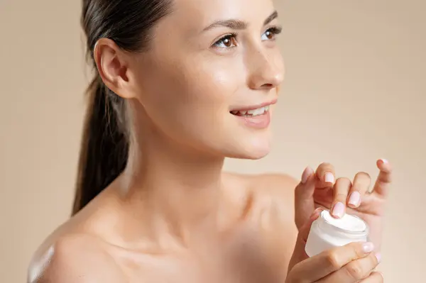 Happy young woman applying cream on face over beige studio background. Pretty brunette with bare shoulders using cosmetic for moisturizing skin. Beauty procedures and spa concept.