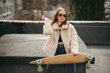 Modaya uygun gözlüklü güzel bir bayan güneş ışığında kaykayıyla çatıda oturuyor. Günlük giysiler içinde beyaz kadın hipster yorucu spor aktivitelerinden dinleniyor..