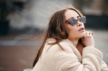 Modaya uygun gözlükler ve sahte kürk mantoyla çatıda poz veren çekici genç bir kadın. Hafif rüzgar onun kısa kahverengi saçlarıyla oynuyor..