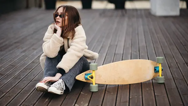 身穿太阳镜 皮大衣和深色牛仔裤的白人女性嬉皮士坐在她滑板附近的屋顶上 年轻黑发女人最喜欢的极端嗜好 — 图库照片