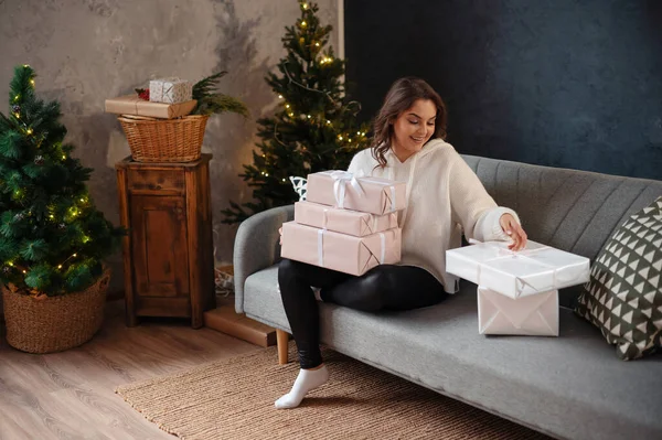 Kıvırcık saçlı mutlu genç bayan Noel ağacının yanındaki kanepede otururken hediyelerini inceliyor. Yeni yıl kavramı