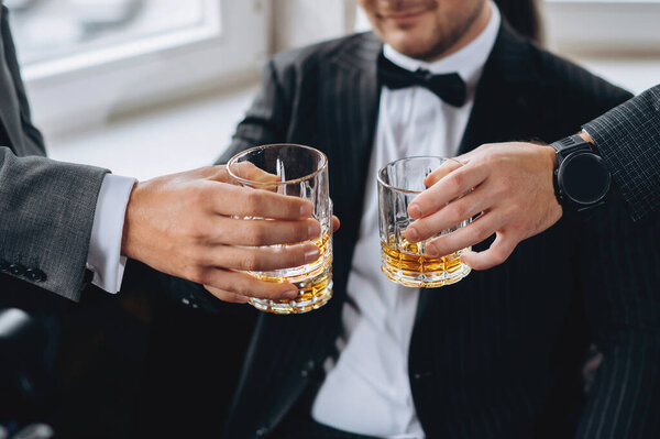 Cropped shot of groom and his friends wear suits holding glasses of whiskey. Wedding day. No face shoot