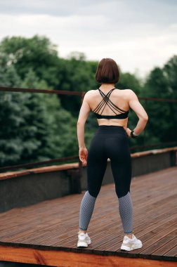 Tahta teras ya da balkondaki Avrupalı sporcunun arka görüntüsü. Genç ve güzel bir kız spor giyer, spor ayakkabı giyer ve bir yerlere bakar. Açık havada spor anlayışı. Yaz bulutlu günü