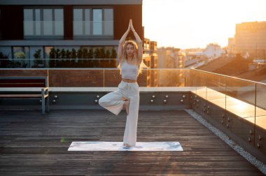 Uzun kıvırcık saçlı genç bir kadın tek bacağının yanında duruyor ve gün doğumunda bir binanın terasında yoga yapıyor.